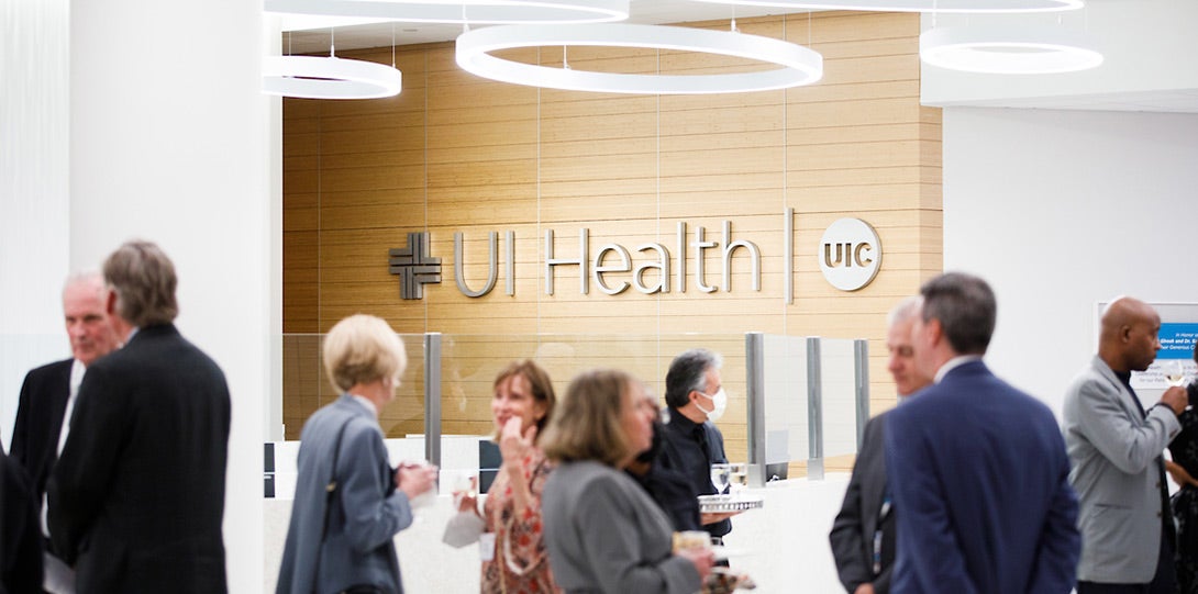 Health leadership and Board of Trustees leadership enjoying a reception, with the info desk and large UI Health logo in the background