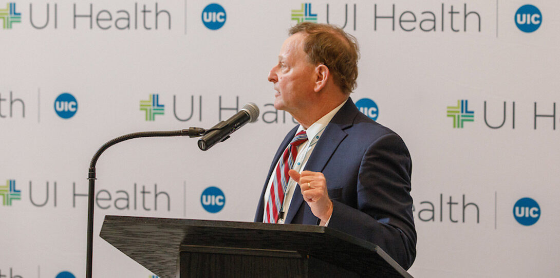 Dr. Barish speaking form behind a podium, standing in front of a backdrop of UI Health logos