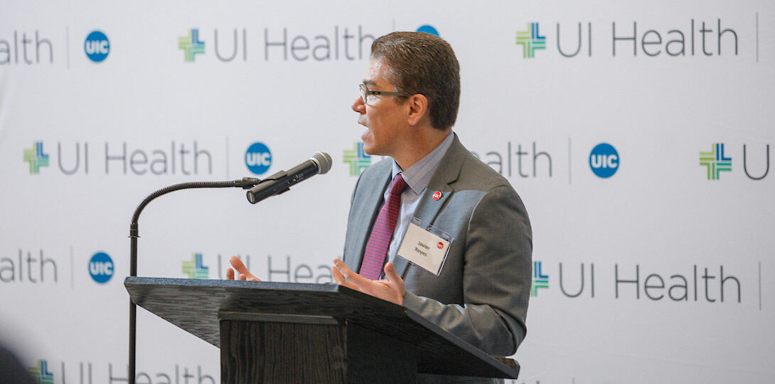 UIC Interim Chancellor Javier Reyes speaking form behind a podium, standing in front of a backdrop of UI Health logos