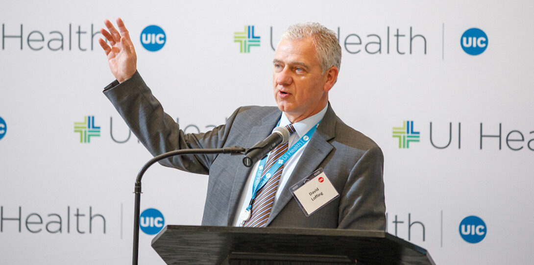 David Loffing speaking into a microphone from behind a podium, in front of a backdrop of UI Health logos
