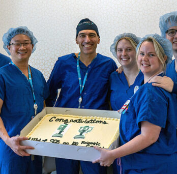 surgeons in blue scrubs posing with a large sheet cake decorated with 