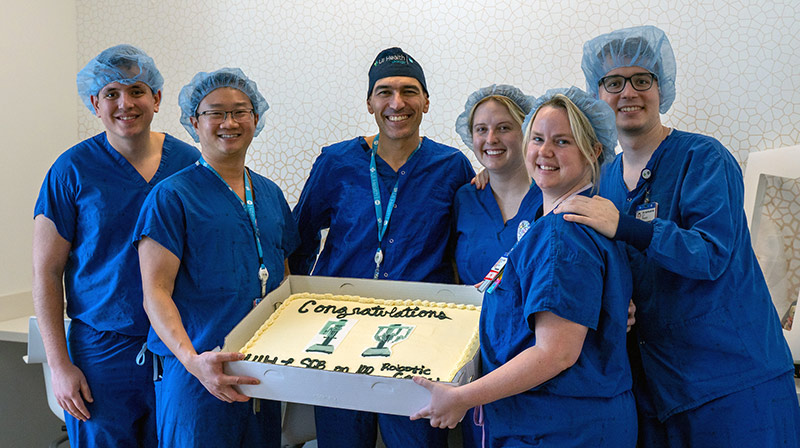 surgeons in blue scrubs posing with a large sheet cake decorated with 