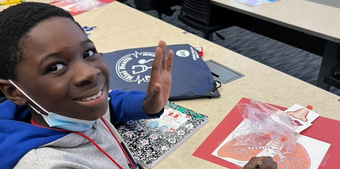 An African American boy sitting at a desk and looking into the camera waving hello