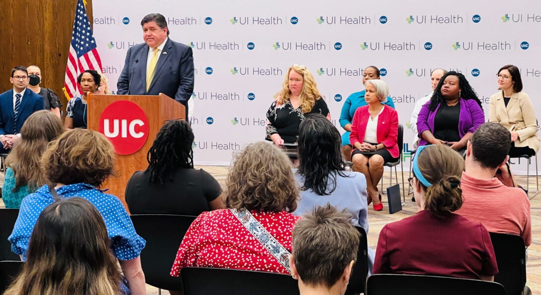 the stage of the press conference with the various speakers sitting in chairs in a row and the governor standing behind a podium, all against a backdrop showing the UI Health logo