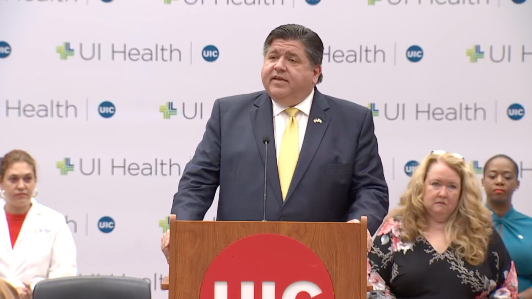 Illinois Governor Pritzker standing behind a podium with a large UIC logo on the front