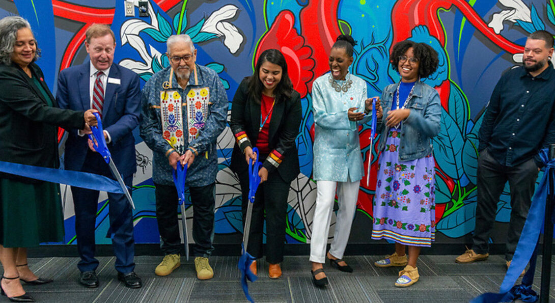 UIC and community leaders and stakeholders standing together and cutting a large blue ribbon front of a colorful mural