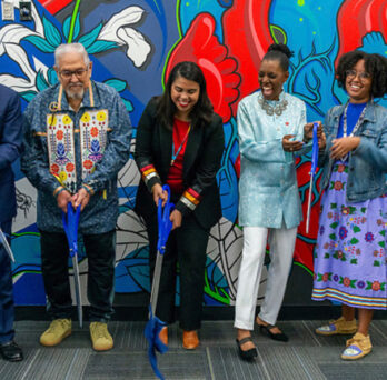 UIC and community leaders and stakeholders standing together and cutting a large blue ribbon front of a colorful mural 