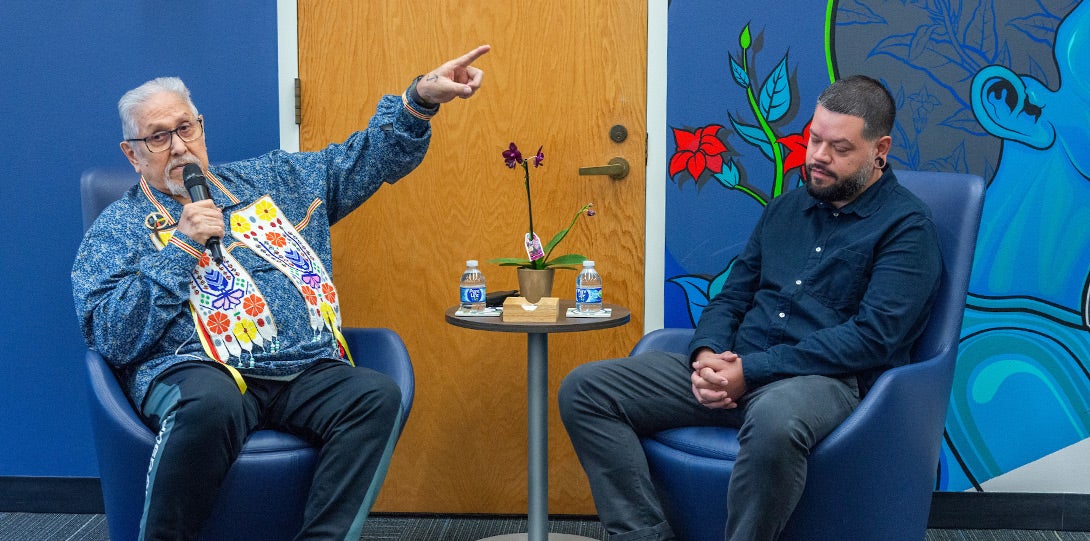 a photo of an older Native American man (the linguist) gesturing toward the mural and speaking into a microphone while a younger man (the muralist) listens