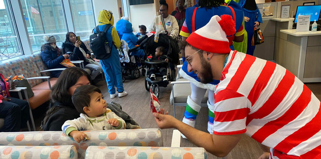 A man dressed as Waldo handing a treat to a latino baby