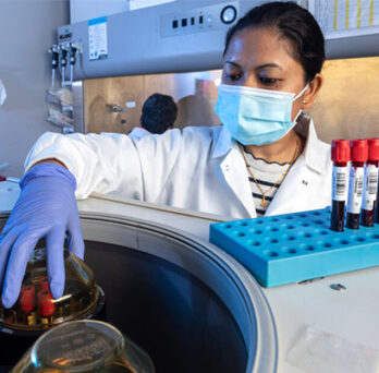 woman in white lab coat and mask removing vials of blood from a centrifuge 