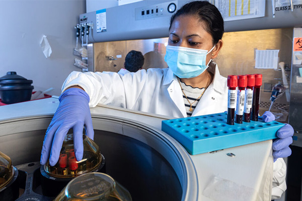 woman in white lab coat and mask removing vials of blood from a centrifuge