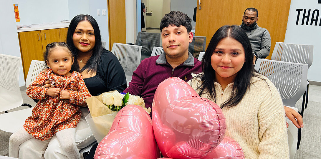 latinx family with a motehr holding a young girl on her lap and a young woman holding several pink heart balloons