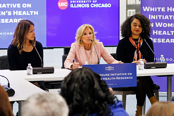 Halle Berry, First Lady Dr. Jill Biden and Alexandra Paget Blanc seated next to each other around a table with video screen and banners in the background