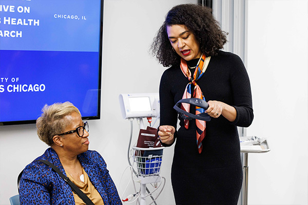 two African American women, one seated wearing a blue jacket, and one standing holding a small electronic device
