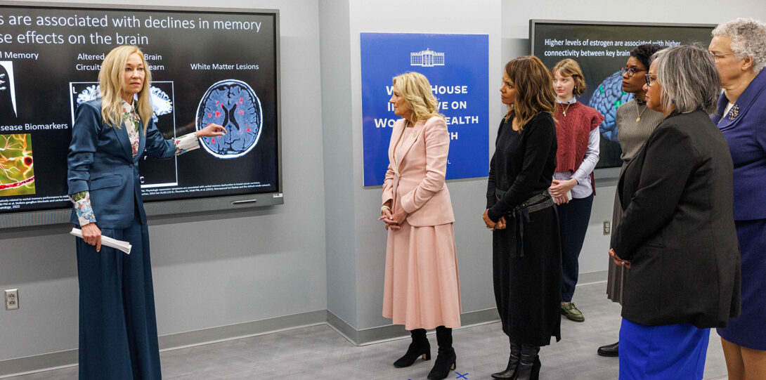 blonde woman in a turquoise suit gesturing to a display of a brain scan as the named people stand to the side, listening