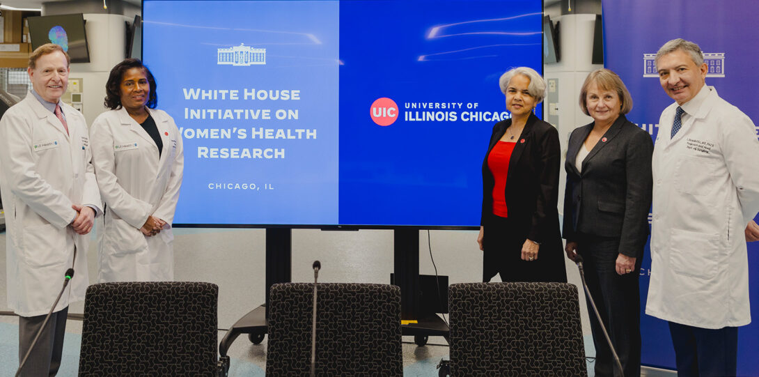 the named peopel posing in a small group next to a large video display which reads White House Initiative on Women’s Health Research