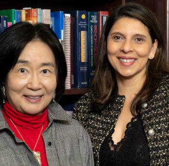 an Asian women with shoulder length black hear, wearing a red turtleneck and a gray jacket, next to a woman long brown hair wearing a patterned gray jacket with black blouse, both standing in front of book shelves 