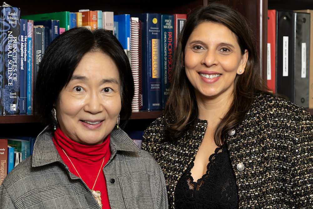 an Asian women with shoulder length black hear, wearing a red turtleneck and a gray jacket, next to a woman long brown hair wearing a patterned gray jacket with black blouse, both standing in front of book shelves