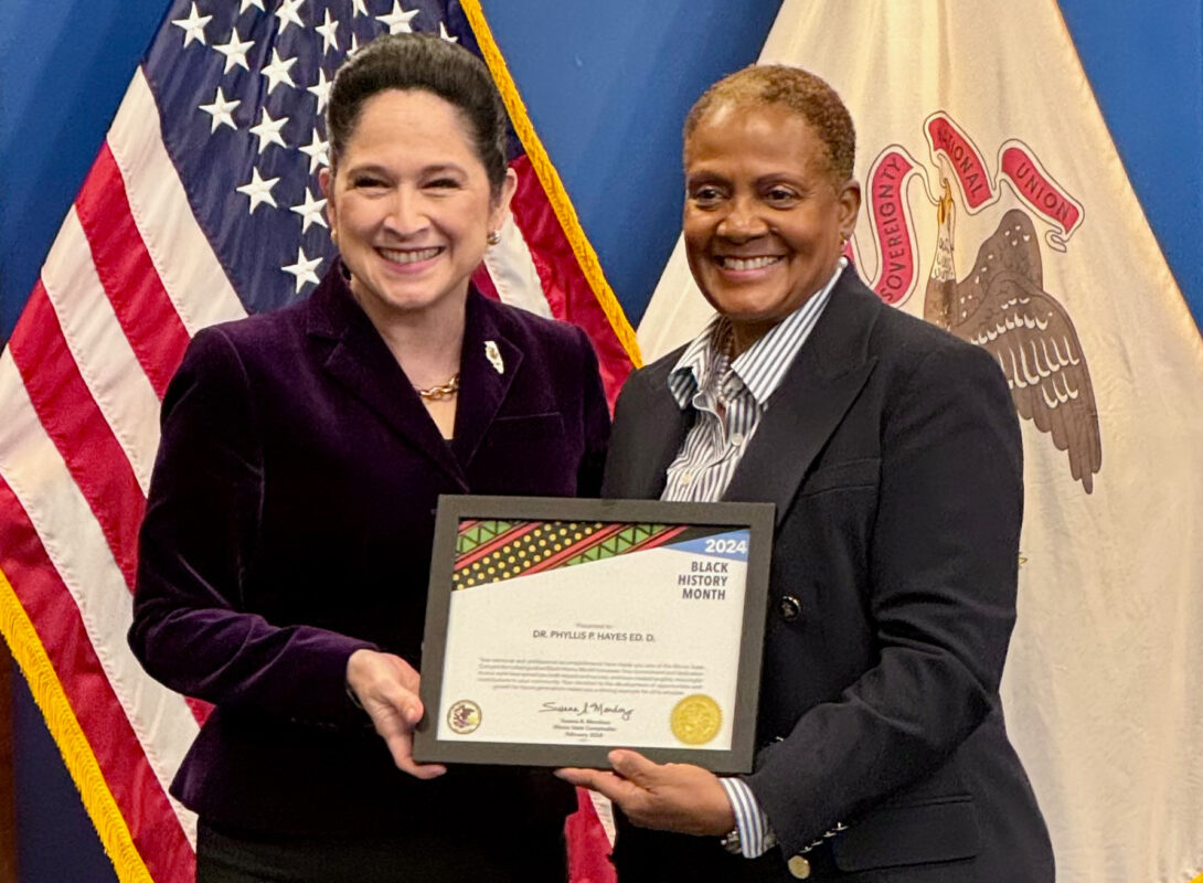 Illinois State Comptroller Susan Mendoza standing next to Dr. Phyllis Hayes, each holding the award certificate, with a U.S. flag and Illinois State flag in the background