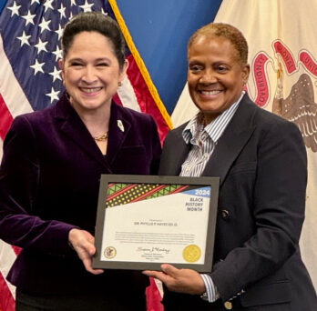 Illinois State Comptroller Susan Mendoza standing next to Dr. Phyllis Hayes, each holding the award certificate, with a U.S. flag and Illinois State flag in the background 