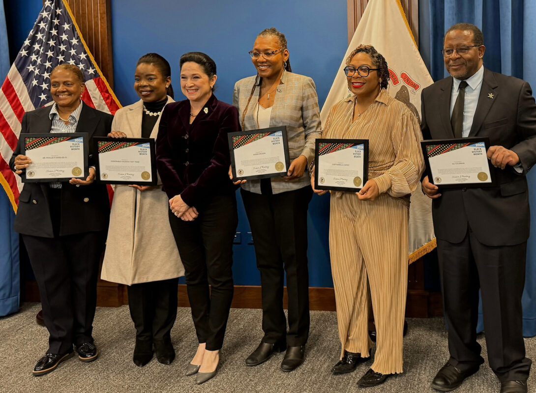 five Chicago African American awardees holding their award certificates and posing with Comptroller Mendoza, with a U.S. flag and an Illinois State flag
