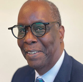 photo of African American man wearing glasses and a blue suit coat, white shirt and blue neck tie 