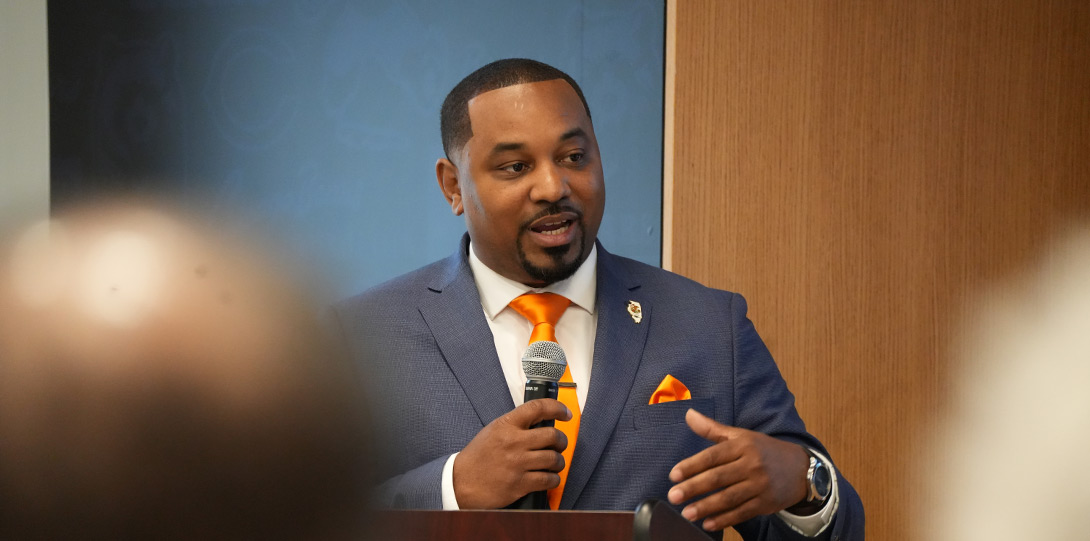 an African American man in a blue suit and orange neck tie, holding a microphone as he is addressing the audience