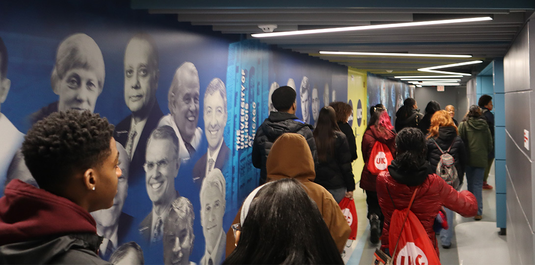 a large group of diverse high schools students in winter attire and wearing bright red UIC backpacks walking down a brightly lit and colorfully decorated hallway in the robotic surgery lab