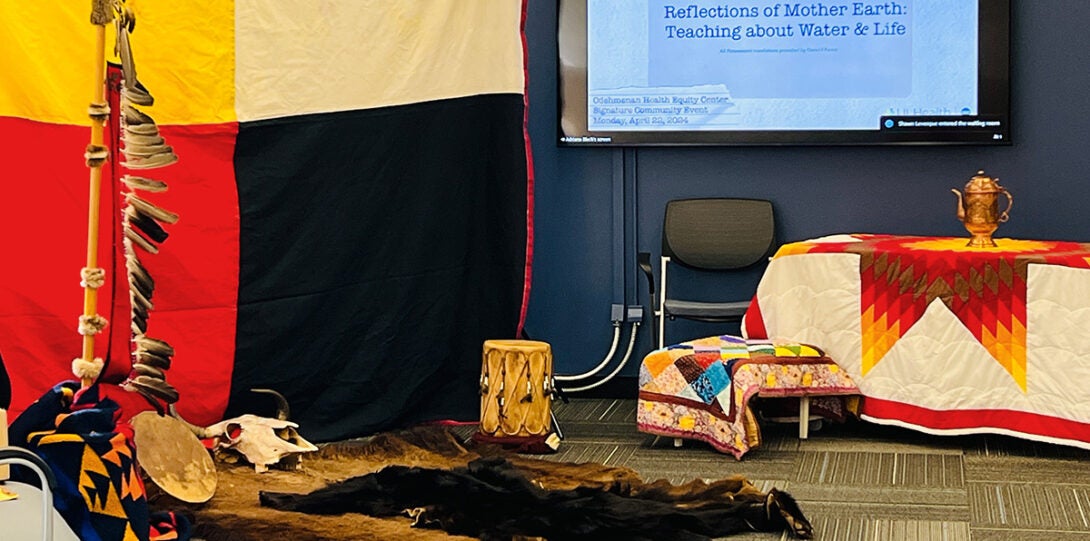 Odeh Center prepared for the presentations with a large blanket with quadrants of yellow, white, red and black, in front of which is an eagle feather staff. On the floor are traditional blankets, various drums, and a buffalo skull and bear hide. There are two tables covered with native blankets, with the larger table bearing a copper carafe.