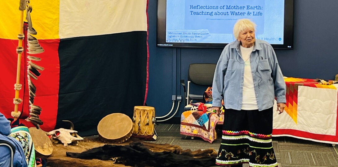 An older native woman with short white hair wearing a traditional ribbon skirt with a white t-shirt and a denim shirt, standing in front of the presentation of ceremonial items.