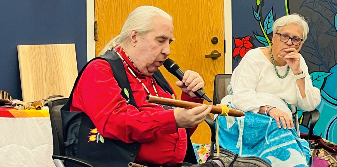 Native man with long white hair in a ponytail, wearing a bright red shirt, black vest and native beads, seated and holding a wooden flute. At right is a woman with short white hair wearing a white blouse, patterned blue skirt and glasses.