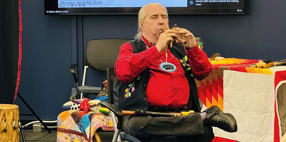Native man with long white hair in a ponytail, wearing a bright red shirt, black vest and beaded necklace, seated and playing a wooden flute with his eyes closed.
