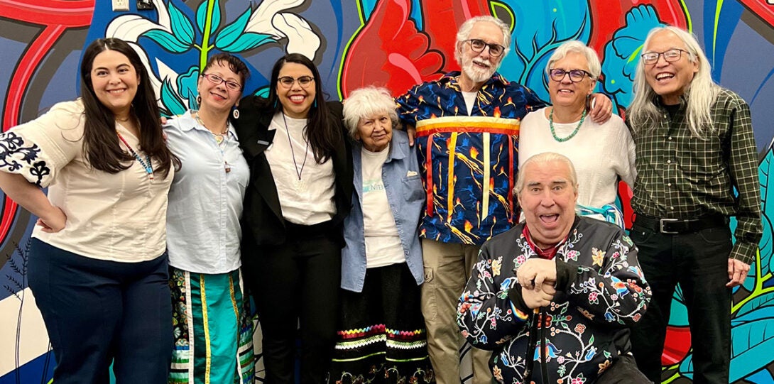 the named people standing in a row with arms around each other’s shoulders in front of a brightly colored mural with native and healing motifs.