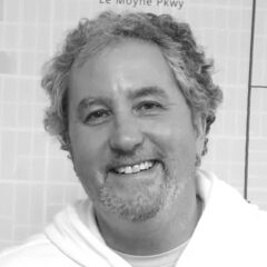 black and white photo of person with curly hair and beard and mustache, wearing a white hoodie and smiling into the camera
