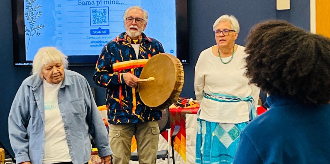 ttendees standing in a semicircle facing the three speakers, who are singing while one of them is playing a native drum.
