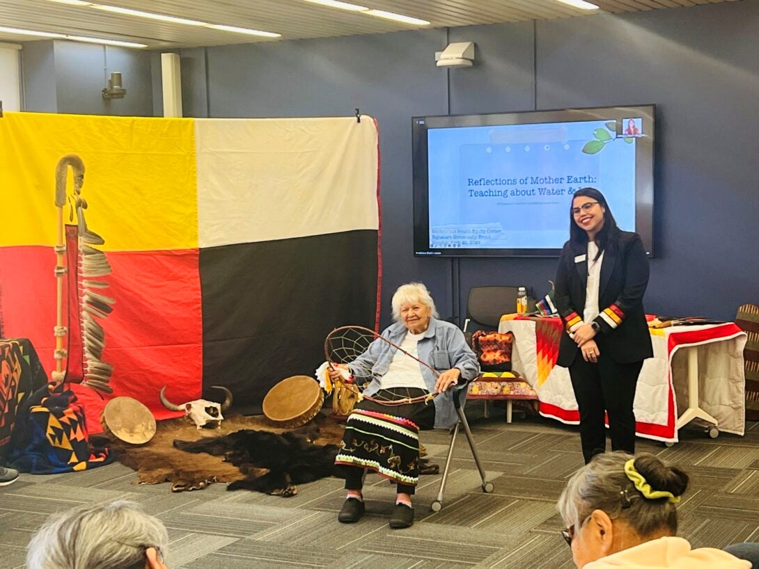 An older Native American woman, wearing a traditional ribbon skirt with a white t-shirt and a denim shirt, is seated and holding a dreamcatcher. Standing next ext to her is a younger woman wearing a black suite. In teh background is a TV screen showing the text 
