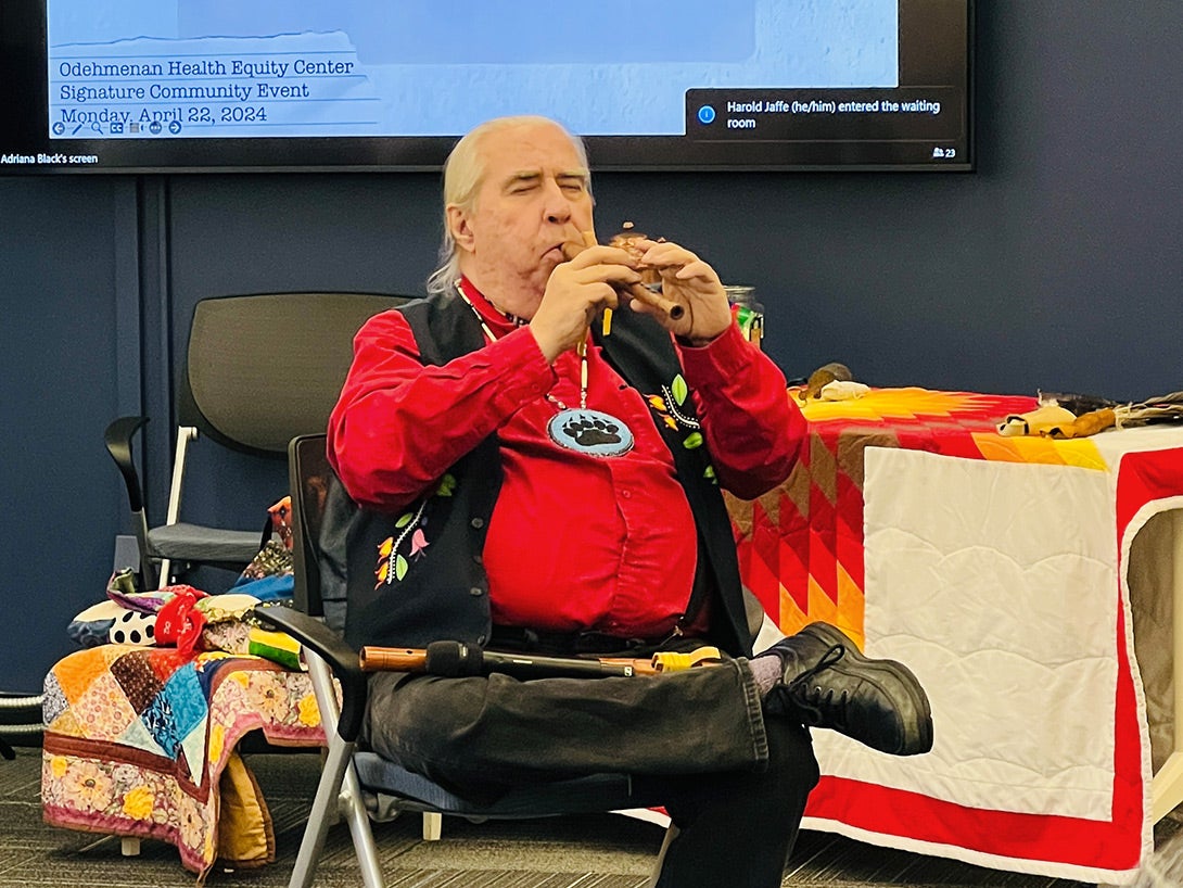 Native man with long white hair in a ponytail, wearing a bright red shirt, black vest and beaded necklace, seated and playing a wooden flute with his eyes closed.