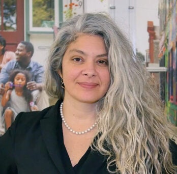 screen grab from the video showing Yamilé Molina wearing a black suit jacket and a necklace, with long salt and pepper hair draped over one shoulder, smiling and standing in front of a colorful mural showing scenes of community life 