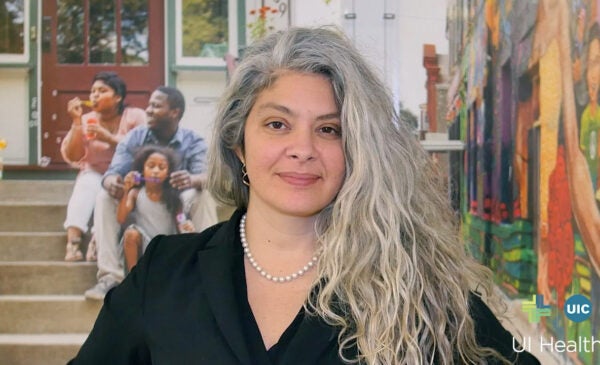Yamilé Molina wearing a black suit jacket and a necklace, with long salt and pepper hair draped over one shoulder, smiling and standing in front of a colorful mural showing scenes of community life