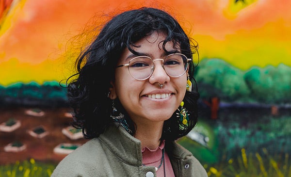 young woman with shoulder-length dark hair, wearing glasses, long earrings and a moss green jacket, smiling into the camera