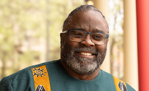 African American man with short salt and pepper hair and a bear and mustache, wearing glasses and a green and yellow jacket with African designs, smiling into the camera