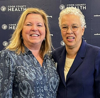 at left a woman with shoulder-length blonde hair wearing a patterned gray blouse, and at right an African American woman with white hair and wearing a white blouse under a blue jacket, both smiling and posing in front of a background with Cook County Health logos 