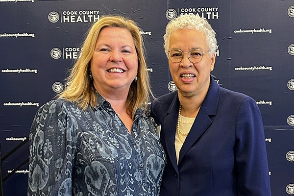 at left a woman with shoulder-length blonde hair wearing a patterned gray blouse, and at right an African American woman with white hair and wearing a white blouse under a blue jacket, both smiling and posing in front of a background with Cook County Health logos