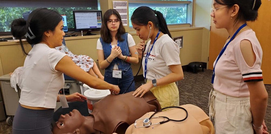 Three female high school students are working with a mannequin in the simulation lab, with one of the medical student mentors looking on.