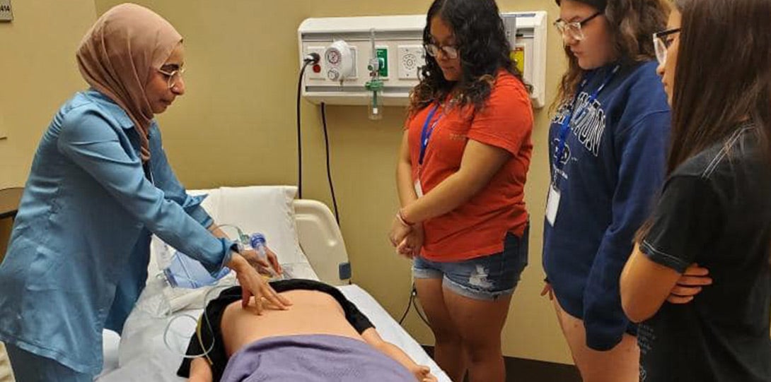 A mentor demonstrating CPR on a mannequin which is lying on a hospital bed in the simulation lab, as three female high school student watch and learn.