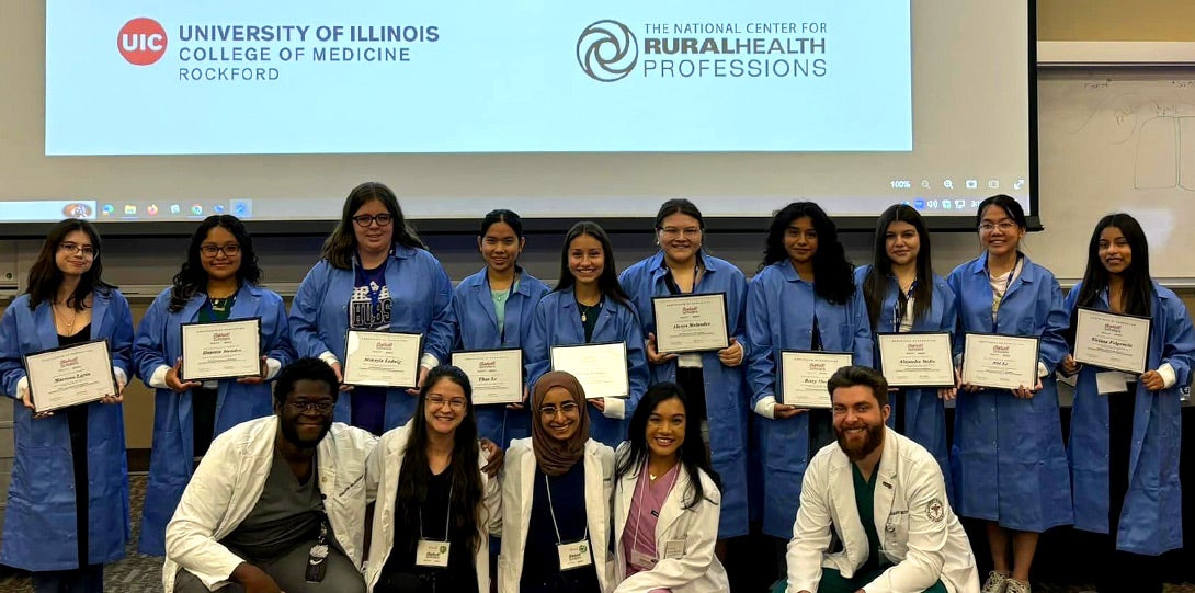 A diverse group of high school students adorned in their blue lab copats and proudly holding their certifiate, with five medical student mentors posing with them, in fron of a screen showing the Univerity of Illinois College of Medicine logo and the National Center for Rural Professions logo.