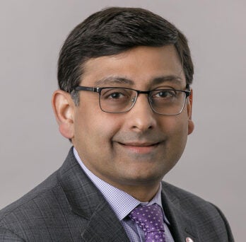 man with short dark hair parted on one side, wearing glasses and a dark gray suit coat, a light colored shirt and purple necktie, smiling into the camera 