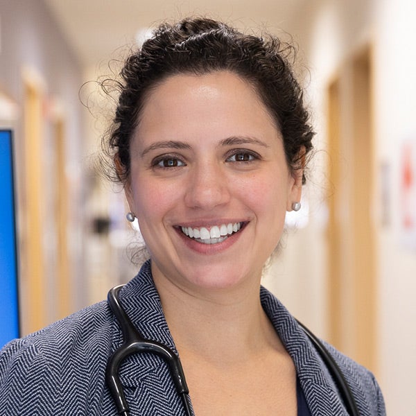 A woman with dark wavy hair, wearing a grey herring bone jacket with a stethoscope draped on her neck, standing in a clinical setting