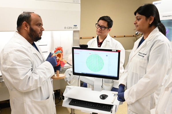 Three people in a laboratory gathered around a 3d bioprinting machine and engaged in discussion