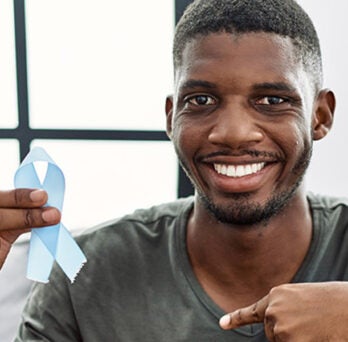 African American man wearing a moss green tee shirt and smiling as he holds up a blue ribbon which represents surviving prostate cancer 